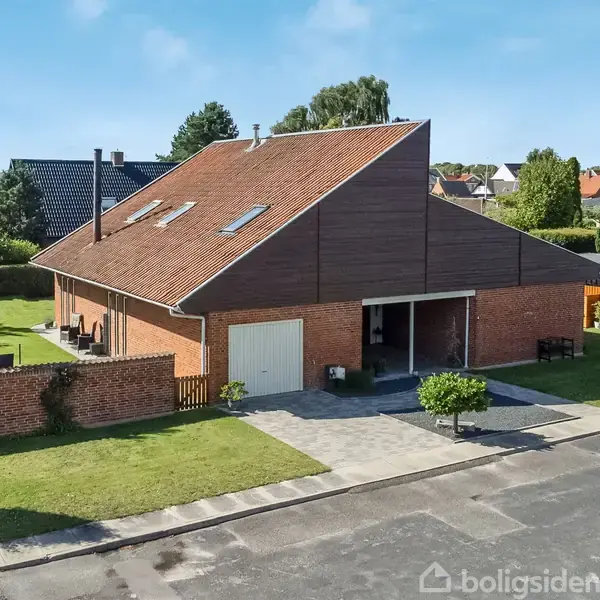 Huset er en stor, rød murstensbygning med et skråt tag og garage. Det står på en velplejet græsplæne i et boligkvarter med andre huse i baggrunden.