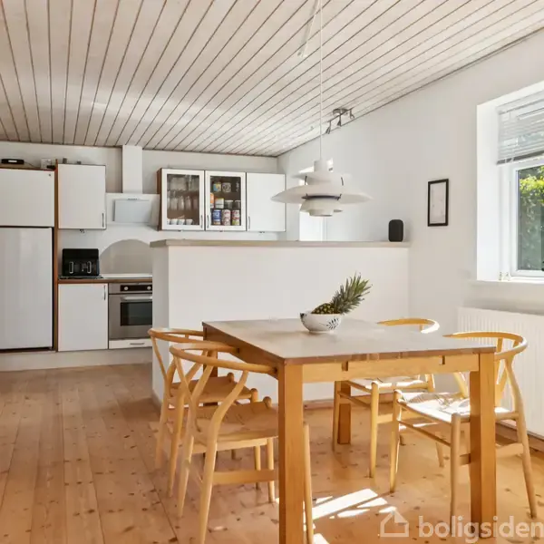 Et lyst træbord med fire stole står i midten af et køkken alrum. En hvid køkkenkrog med ovn og skab er i baggrunden. Et vindue lader dagslys komme ind.