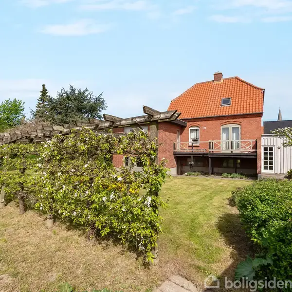 Rødt murstenshus med orange tegltag står i en have med grønne planter og græsplæne. Der er en terrasse og hegn af blomstrende buske.