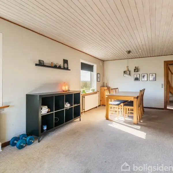 Stue med spisebord og stole, oplyst af vinduer med blå gardiner. På væggen hænger billeder, og der er hylder med dekorationer. En dør fører til en trappe.