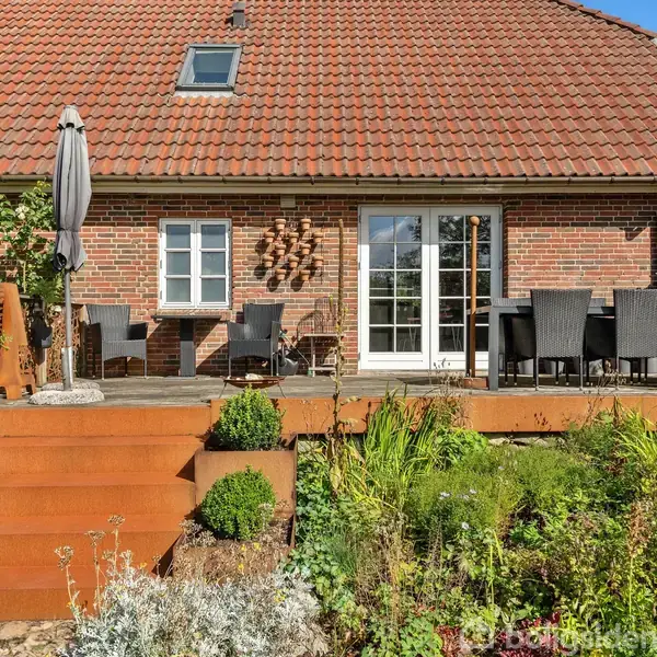 Rødt murstenshus med høj terrasse, hvorpå der står sorte havemøbler og en lukket parasol. En trappe fører til en velplejet have med grønne planter. Husnummeret "62" ses.