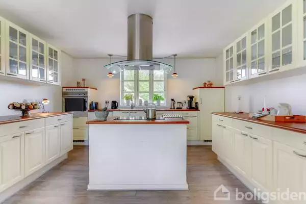 Moderne køkken med hvidt skabsmøblement og centralt placeret kogeø. Over kogeøen hænger en emhætte. Rummet er lyst med et stort vindue og trælaminatgulv. Intet synligt tekst.