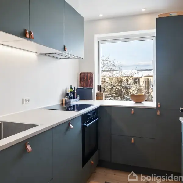 Et moderne køkken med grå skabe og hvide bordplader. Der er en indbygget vask og ovn. Et vindue giver udsigt til et træ. Kaffemaskine står i hjørnet.