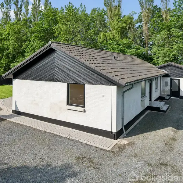 Et hvidt hus med sort tag står på en grusbelagt indkørsel, omgivet af grønne træer og en græsplæne. Ved siden af huset er en sort garage.