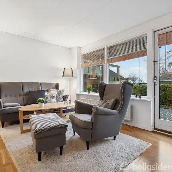 En stue med en grå sofa og lænestol, der står ved et træbord. Store vinduer med udsigt til en terrasse. Mørke lamper står i hjørnerne. Ingen tekst til stede.