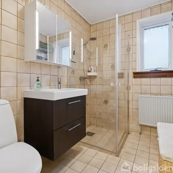 Gråt sofamøblement med sofabord står i en lys stue. Store vinduer giver udsigt til en terrasse og have. To gulvlamper og tæppe skaber en hyggelig atmosfære.