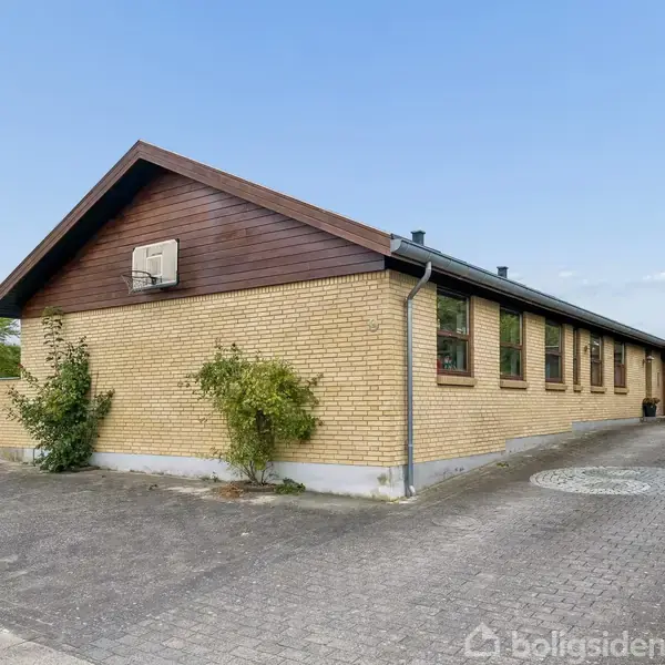 Et gulstenshus har en brun trægavl. Det står på en flisebelagt indkørsel med græs på siden. Der er en garage og luftkonditioneringsenhed på gavlen.