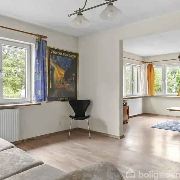 Stue med en sofa ved store vinduer og en plakat af Vincent van Gogh på væggen. Et tilstødende rum med spisebord og gule gardiner kan ses i baggrunden.