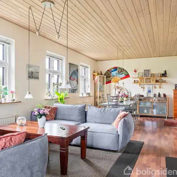 Grå sofaer står omkring et trækaffebord i en lys stue med trægulve. Vinduer til venstre giver naturligt lys. Spisebord og skænk i baggrunden.