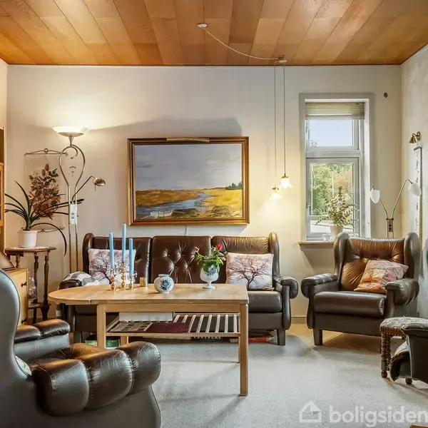 Stue med lædersofa og lænestole omkring et træbord, dekoreret med stearinlys og blomster. Kunstmaleri hænger på væggen, omgivet af naturligt lys fra vinduerne. Reol til venstre.