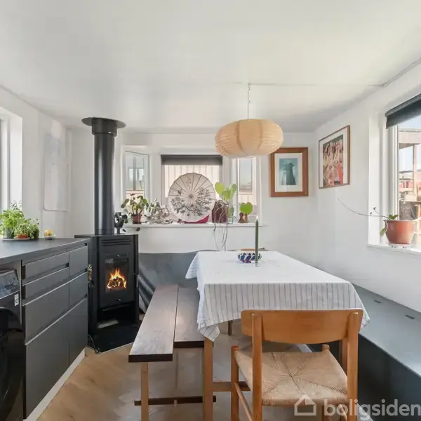 En hyggelig spisestue med et træbord og bænke, hvor der er tændt op i brændeovnen. Omgivet af vinduer med planter, vaskemaskine og indrettet med moderne møbler.
