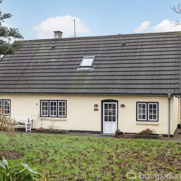 Et hvidt hus med mørkt tag står i en grøn have. Der er flere vinduer og en havedør. Græsset foran huset er delvist dækket af brune blade.