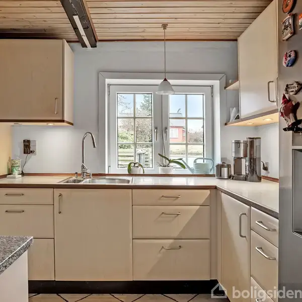 Køkkenet har hvide skabe, en vask ved vinduet og en bordplade. Der hænger en lampe over bordet, og et køleskab med magneter ses til højre.