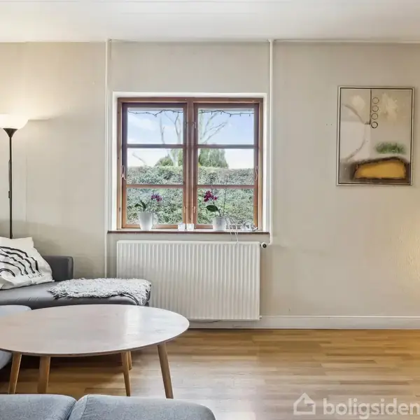 Grå sofa står ved siden af et rundt træbord. Vinduer viser en have udenfor. Væggene har kunstmalerier, gulvet er træ, og stuen er lyst op af gulvlamper.