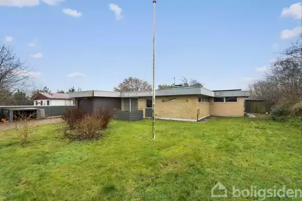 En en-etagers murstensvilla med fladt tag står i en grøn have, omgivet af buske og træer. En flagstang er placeret midt i græsplænen.