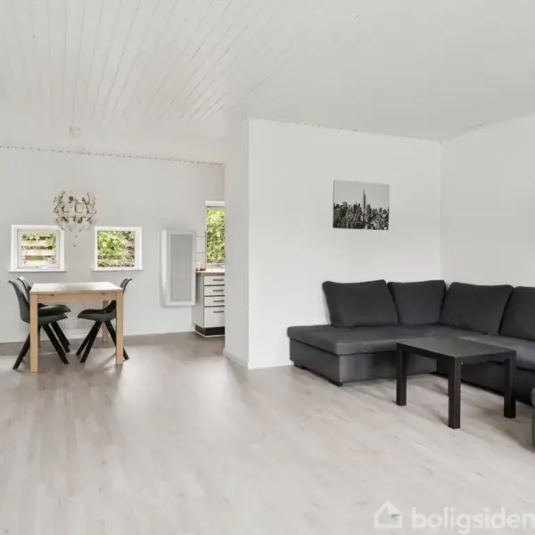 En grå hjørnesofa står i en lys stue ved siden af et sort sofabord. En spisestue med træbord og sorte stole er i baggrunden, understøttet af hvide vægge.