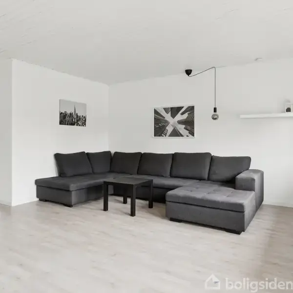 Grå hjørnesofa står stille i et lyst rum med trægulv. Et lille sort bord er foran sofaen. To billeder hænger på væggen, og der er et køkken til venstre.