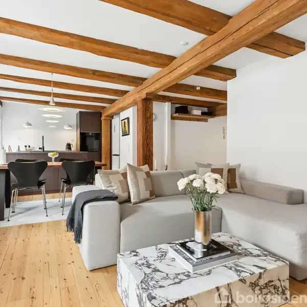 Sofaen står i stuen med et marmorbord foran, der har en blomsterbuket. Køkkenet med spisebord og stole ses i baggrunden. Trævægge og gulv giver en hyggelig stemning.