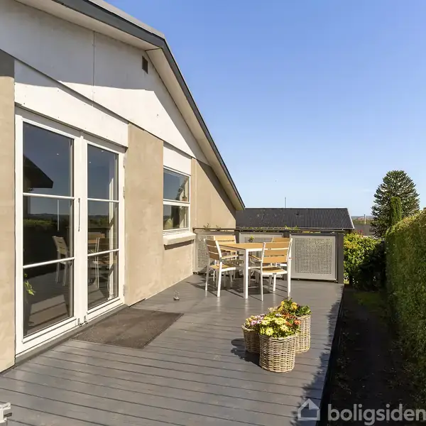 Hus med grå terrasse har havemøbler og blomsterkurv, omkranset af en grøn hæk. Sollys skinner over det moderne, minimalistiske design, hvilket skaber en indbydende udendørs opholdsstemning.