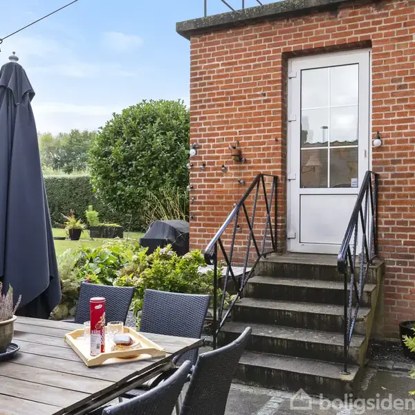 En udendørs terrasse med et bord og stole, hvor der ligger en bakke med blomster og en dåse Pringles. En trappe fører til en dør i en murstensvæg, omgivet af planter.