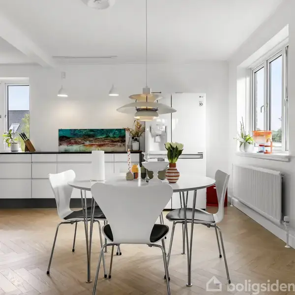 Et rundt spisebord med hvide stole står i et lyst køkken. På bordet er en vase med blomster. Køkkenet har hvide skabe og store vinduer, der giver naturligt lys.