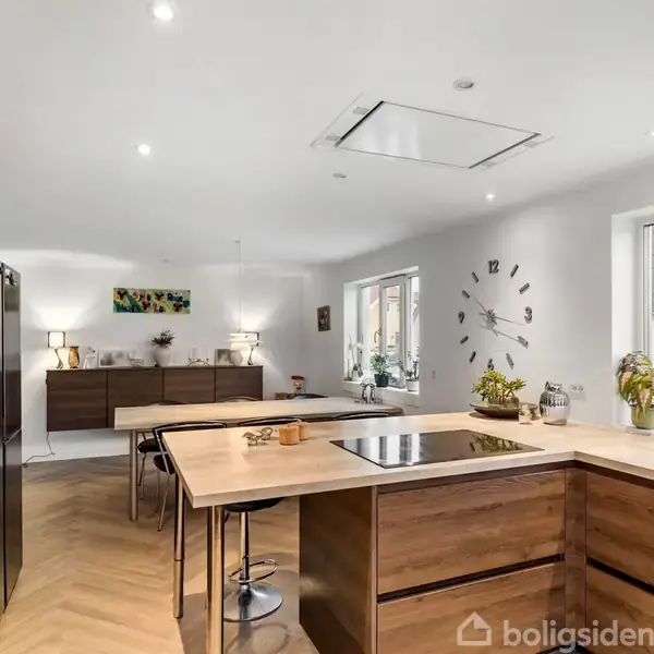 Et moderne køkken med træfinish køkkenø med indbygget kogezone; omgivet af åbent opholdsrum med spisestue. Ur på væggen, planter på bordet og rummelige vinduer i baggrund.