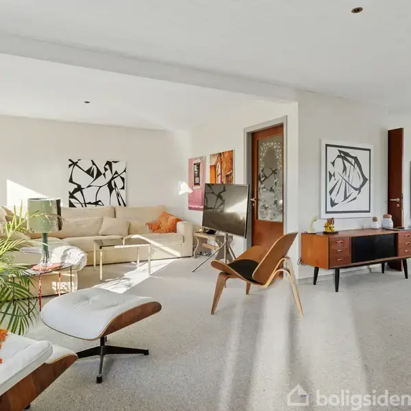 En lys stue med moderne møbler består af en beige sofa, et sofabord og en lænestol med puf. Der er kunst på væggene og planter i hjørnet.