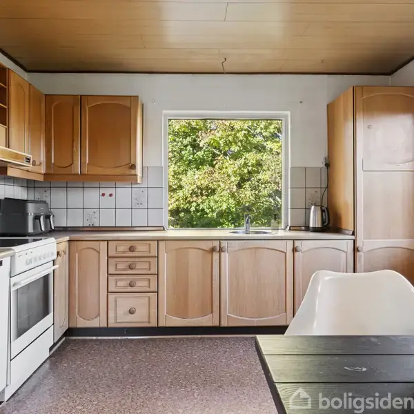 Et køkken med træskabslåger og hvid opvaskemaskine, mikrobølgeovn og elkedel, set ved et vindue ud til grønne træer. I forgrunden står et bord med en skål med frugt.