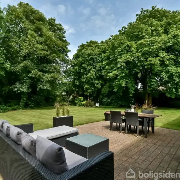 En udendørs terrasse med sofaer og et spisebord står på en flisebelagt overflade. I baggrunden ses en stor græsplæne omgivet af frodige grønne træer.