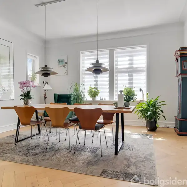 Spisebord med seks stole står på et tæppe i et lyst rum, omgivet af planter og kunst. Store vinduer giver naturligt lys.