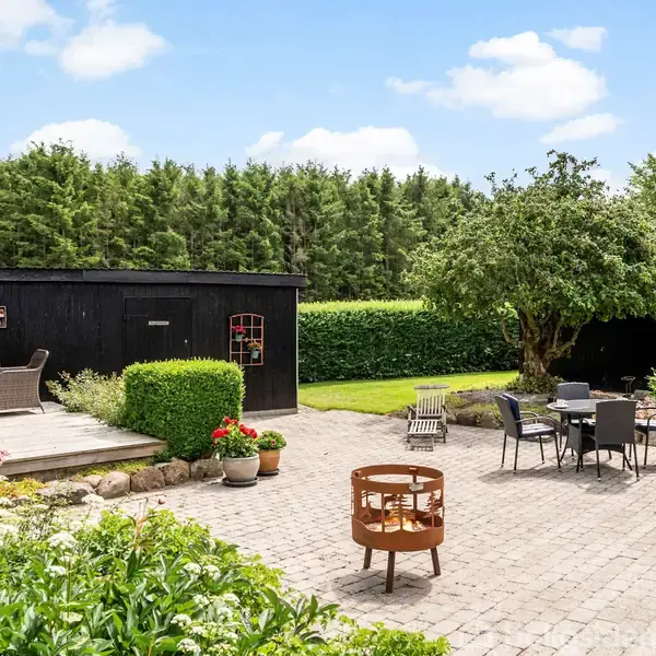 En brolagt have med havemøbler står under et træ. En terrasse med flere møbler er tæt ved. Buske og en tæt hæk omkranser området, med en blå himmel og skyer ovenfor.