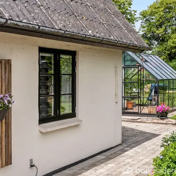 Et hvidt hus med sort vindue har blomster på en trævæg. Til højre ses en lille, glasdrivhus med planter. Solrig have med fliser og grønne buske.