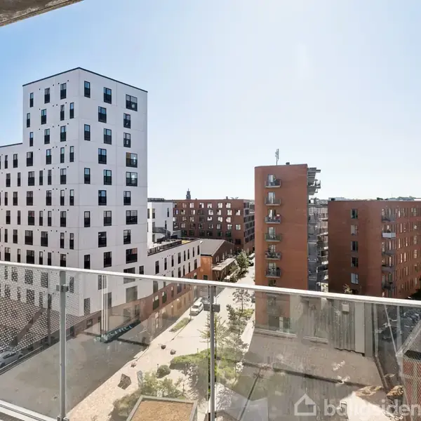 Altan med glasrækværk med udsigt til moderne lejlighedskomplekser. Flere bygninger med varierende højder og farver strækker sig ind i det fjerne under en klar blå himmel.