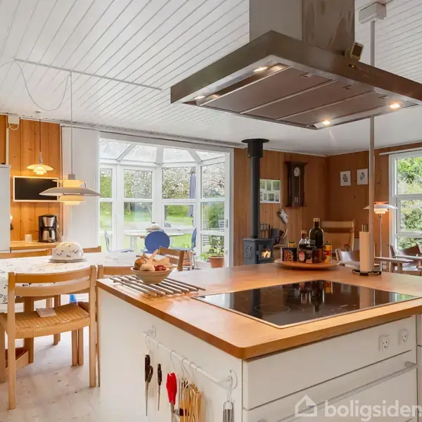 Køkken med kogeø og emhætte omgivet af moderne møbler og spisebord; store vinduer giver udsigt til en grøn have, hvilket skaber en lys og indbydende atmosfære.