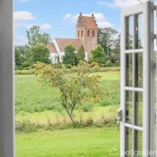 Et smukt landskab set gennem åbne franske døre. En kirke med et rødt murstens tårn står blandt træer. Grønt græs og en blå himmel skaber en fredelig scene.