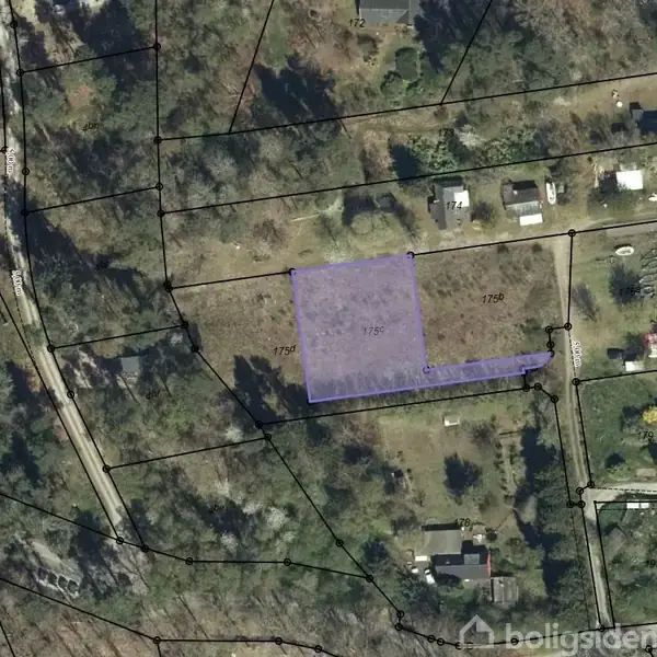 Landsby omgivet af frodig skov ses ovenfra. Flere huse ligger spredt mellem træerne, og i baggrunden er åbne marker med gule afgrøder under en overskyet himmel.