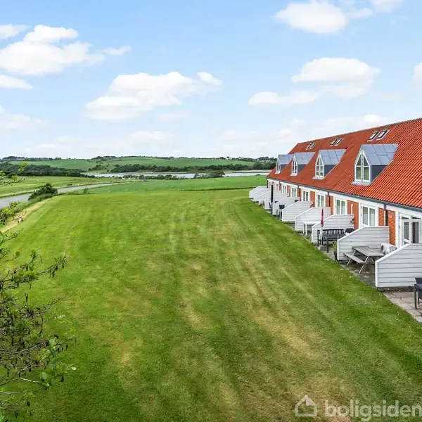 Rækkehusene med røde tage og hvide facader står på en lang række. De grænser op til en stor, grøn græsplæne med udsigt over det kuperede landskab under en delvis skyet himmel.