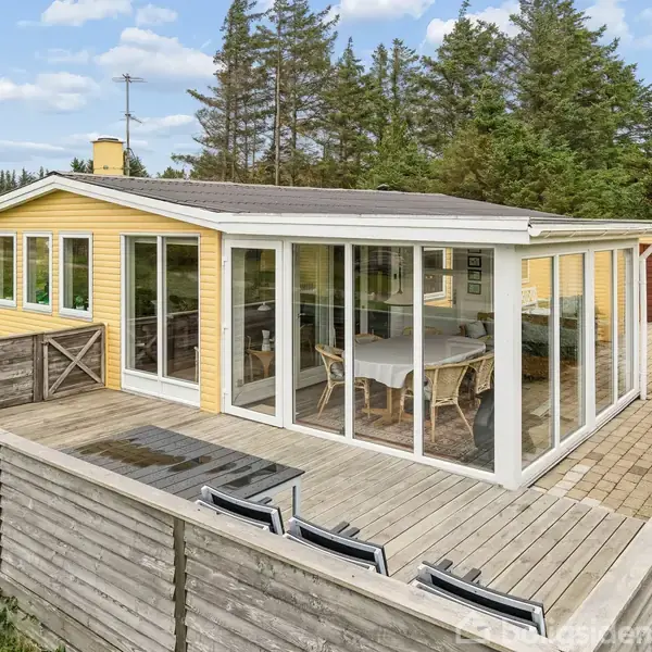 Et gult sommerhus med store vinduer og terrasse. Et bord med stole står i udestuen. Omkranset af træer på en stor græsplæne.