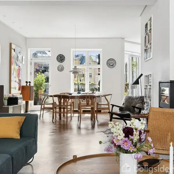 Stue med moderne indretning, hvor en grøn sofa står forrest med en gul pude. Et spisebord ses i baggrunden ved store vinduer. Der er en tændt pejs og blomsterdekorationer.