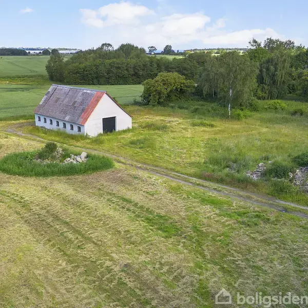 En hvid bygning med rødt tag står stille på en åben mark, omgivet af grønne marker og træer under en blå himmel.