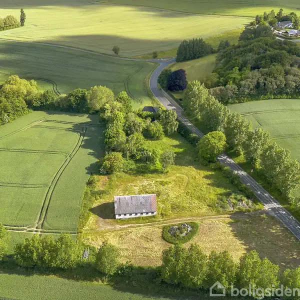 Et hus ligger stille omgivet af marker og træer på en solrig dag. En vej løber gennem landskabet, og små landsbyer kan ses i det fjerne.