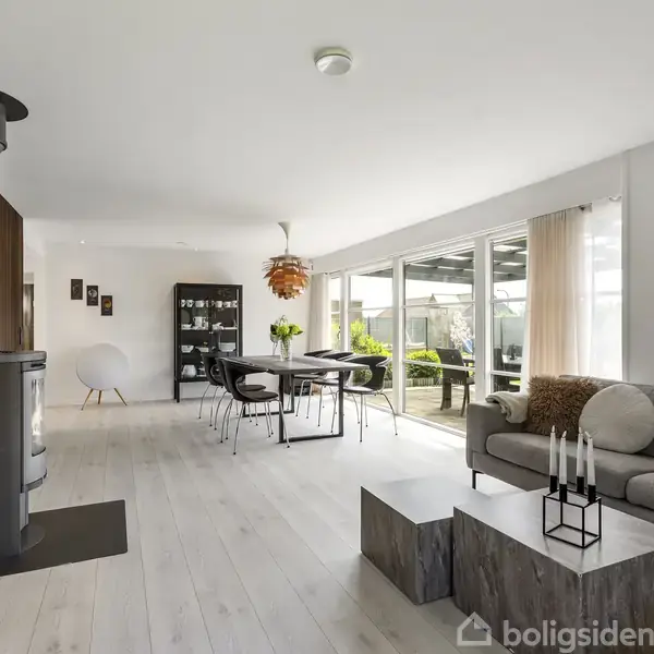 Brændeovn står ved væggen, ikke i brug, i en moderne stue med lyse trægulve. En grå sofa med sofabord, spisebord med sort stole; store vinduer lukker naturligt lys ind.