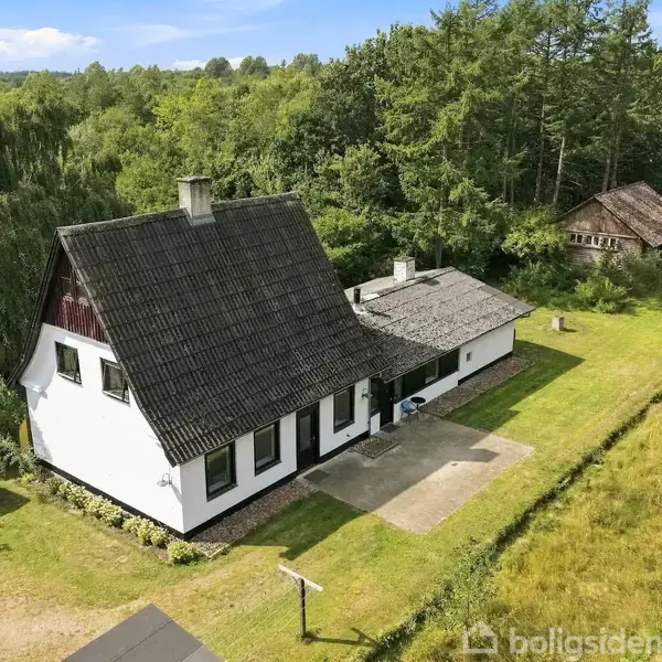 Et hvidt hus med sort tag står i en skovfyldt omgivelser. Foran huset vejer et dansk flag, og til højre ses en mindre træbygning.