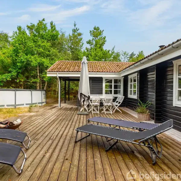 Terrasse med to sorte liggestole og et bord med stole under en parasol; omringet af træer. En mørk træbygning ligger til højre med hvide vinduer.
