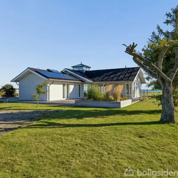Et moderne hus med solpaneler står på en velholdt græsplæne nær vandet. Et stort træ med beskårne grene ses i forgrunden, omgivet af natur og åbne marker.