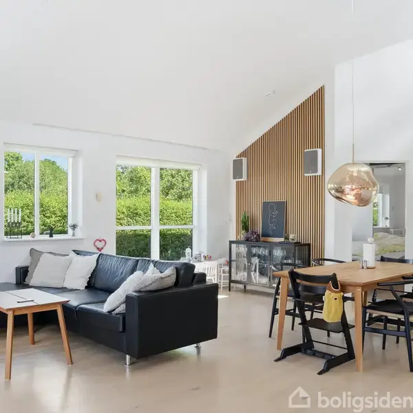 Sort sofa med puder står ved siden af et lille bord i en stue med store vinduer, træbord og stole. De hvide vægge har kunstværker, og rummet er lyst og åbent.