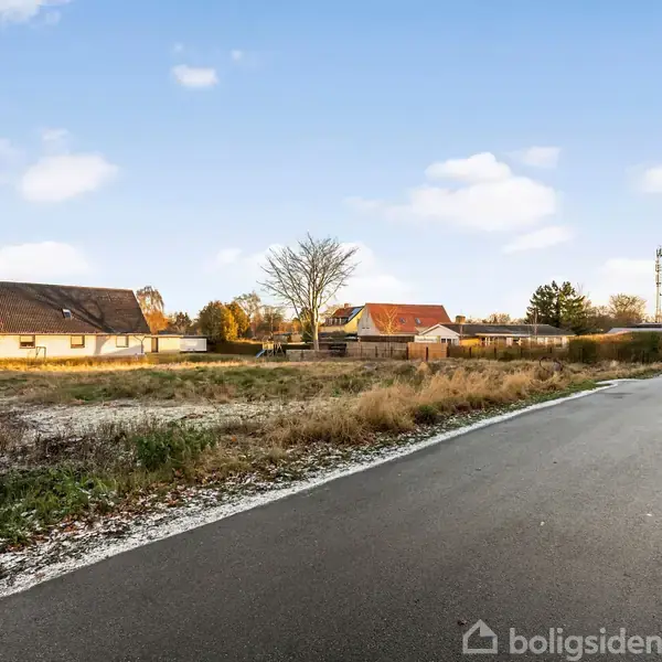 En asfalteret vej løber langs en række huse med store græsarealer på en klar dag. Buske og træer står tilfældigt i et beboelsesområde.