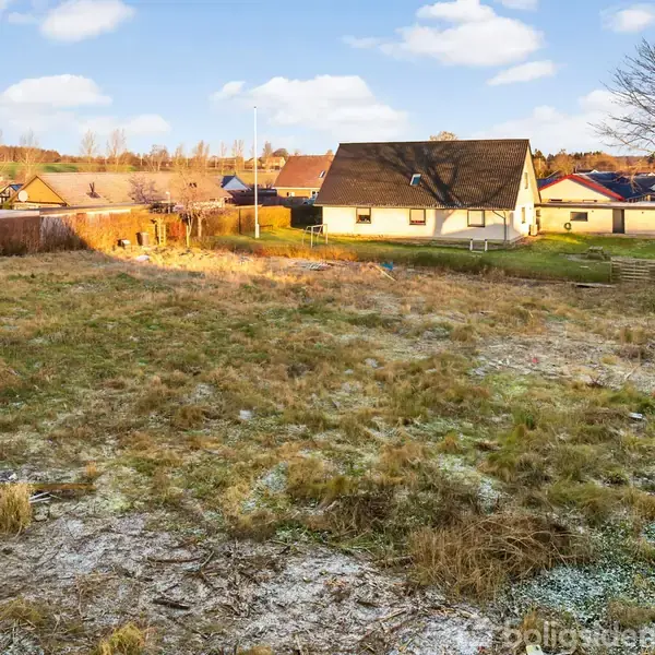 En åben grund ligger stille med ujævn græs og frost, omgivet af en række huse i en villakvarter med blå himmel og spredte skyer.