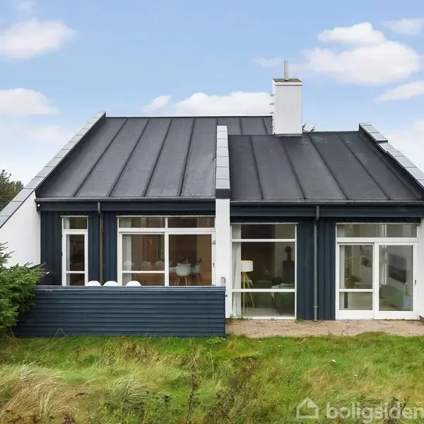 Et moderne hus med sort tag og hvid facade ligger i en græsplæne. Store vinduer ses på forsiden, og til højre er der indgang til en terrasse.