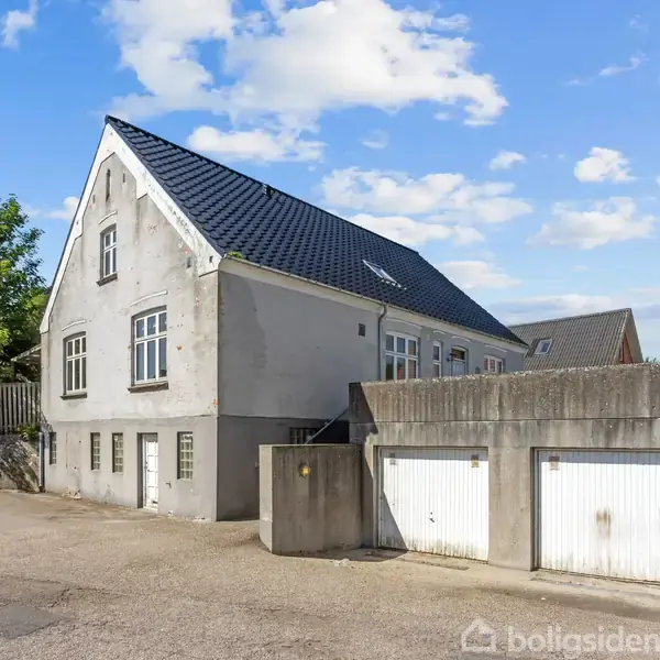 Et stort hvidt byhus med blåtag står stille ved siden af to lukkede garager. Omgivet af en parkeringsplads og grønne træer under en blå og skyet himmel.